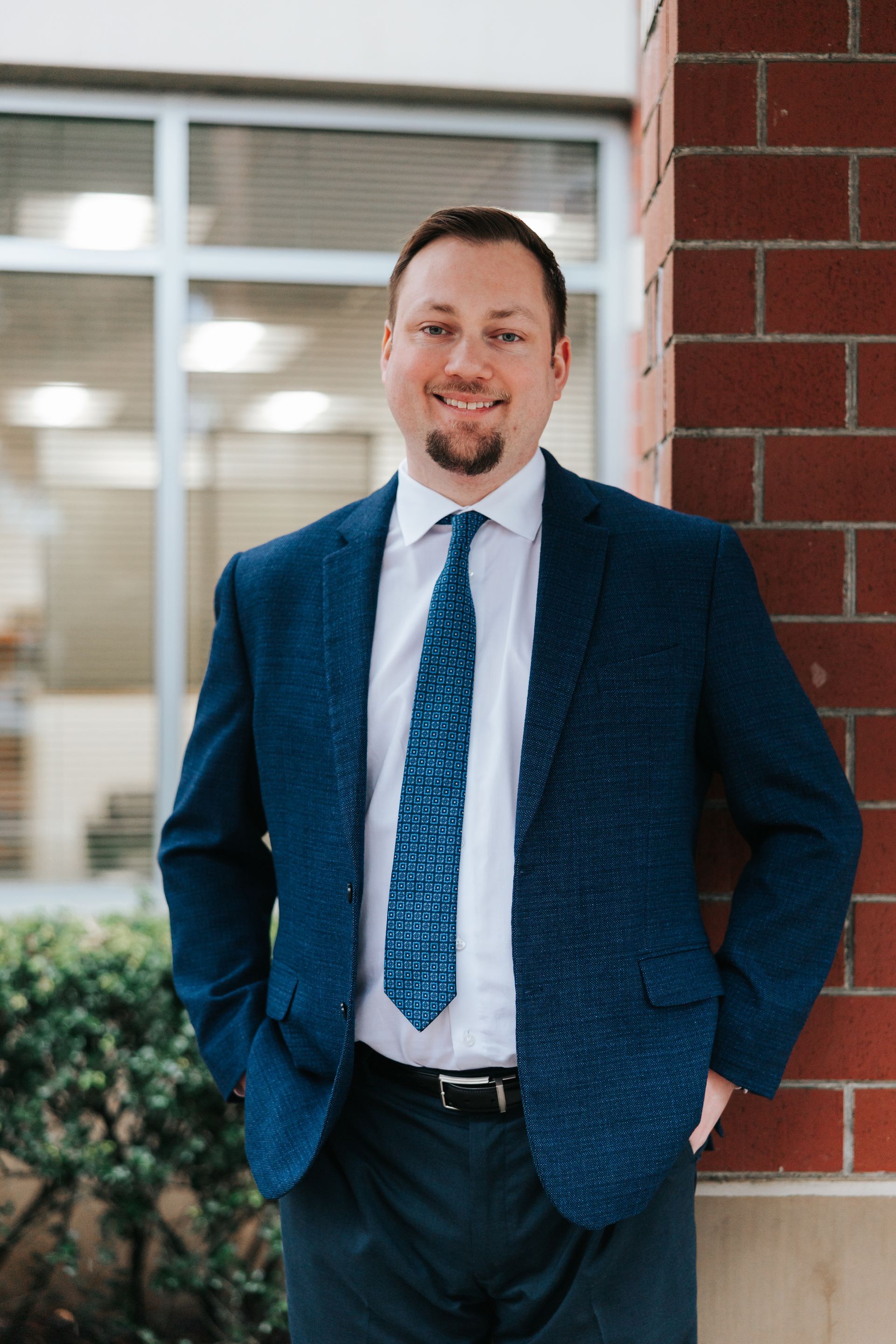 Woman in black suit leans against a brick pillar, hands on hips, smiling, in a corridor.