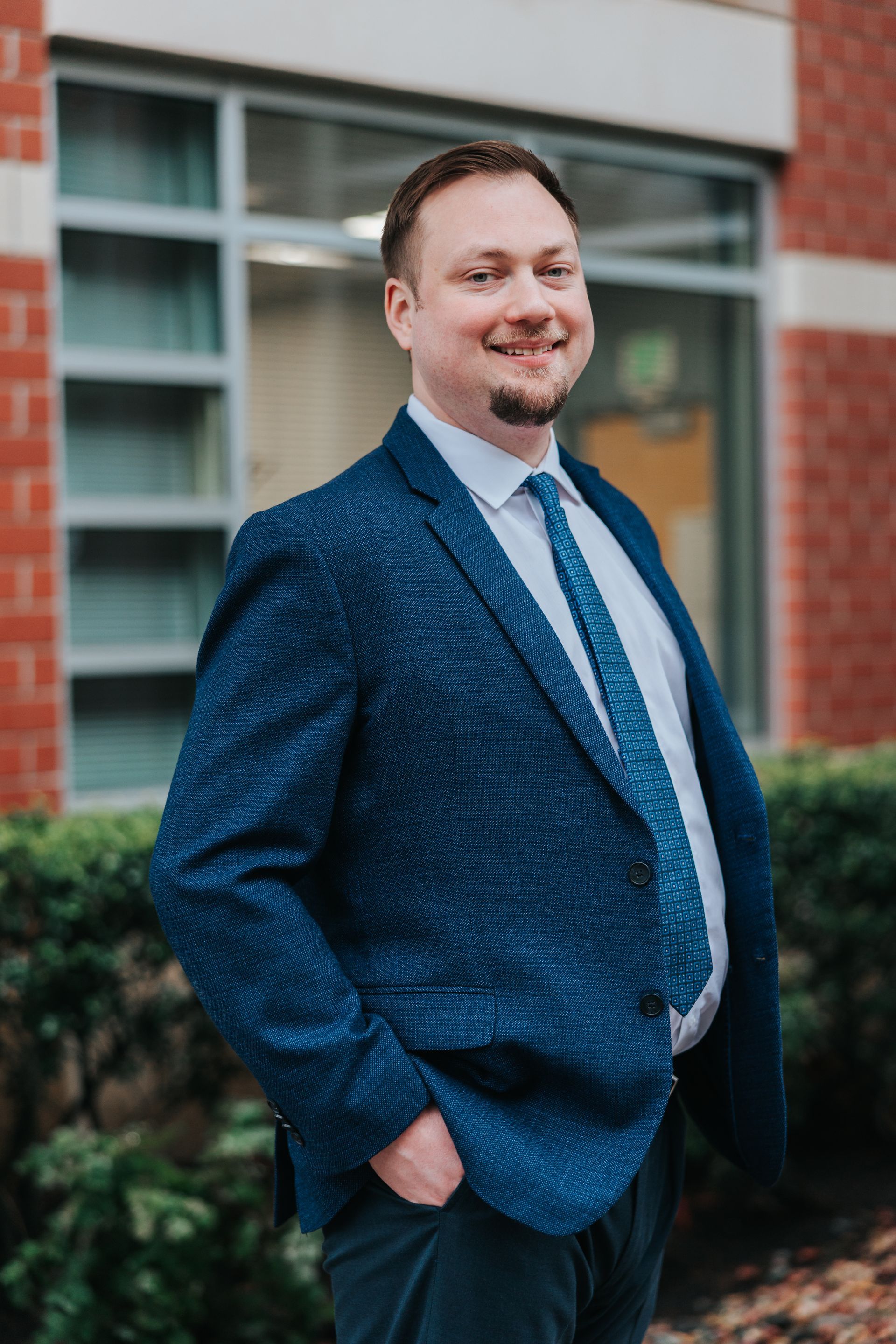 Woman in black suit leans against a brick pillar, hands on hips, smiling, in a corridor.