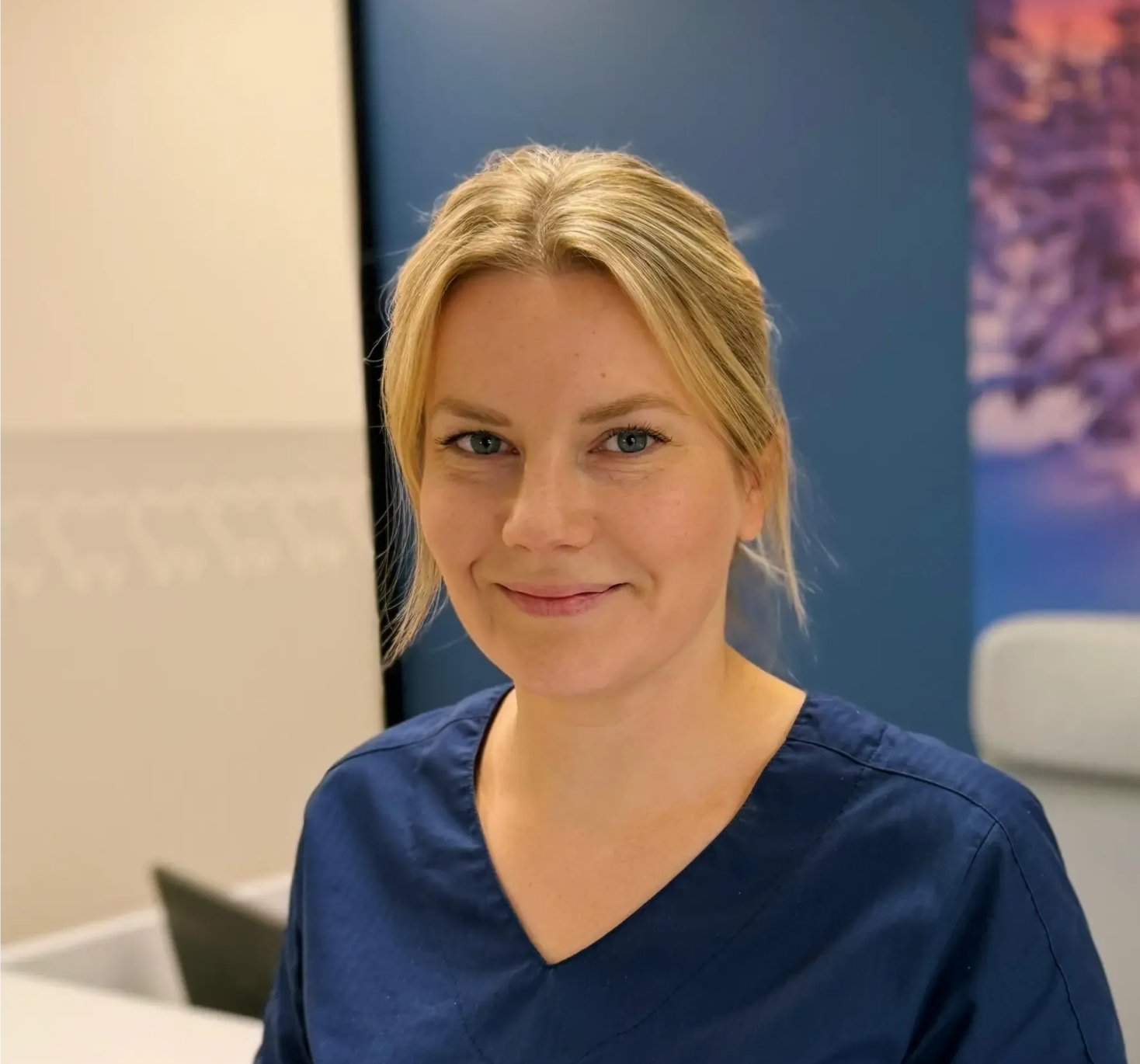 Kvinne med blondt hår iført en marineblå skrubbtopp, smilende på et legekontor.
