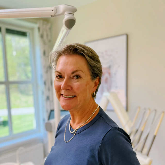 Kvinne i blå skjorte smiler på et tannlegekontor.