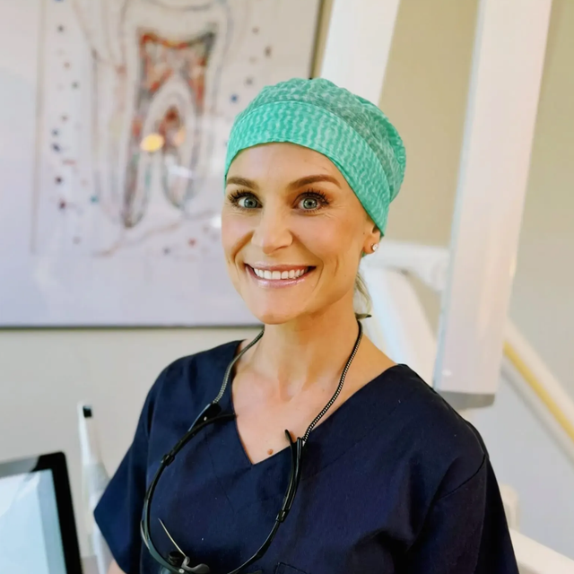 Tannlege smiler, iført skrubbhette og briller, på et tannlegekontor med utstyr og tanndiagram.