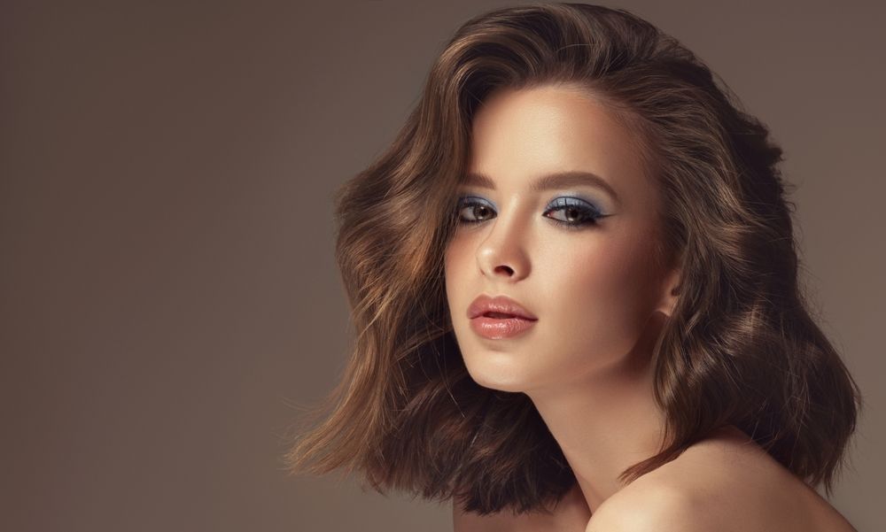 Woman with wavy brown hair, blue eyeshadow, and nude lipstick, looking towards the camera.