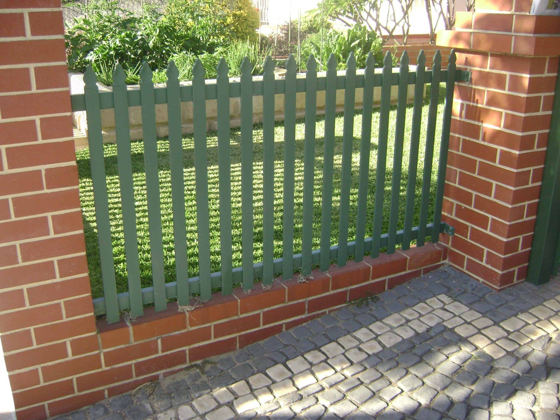 Green picket fence between brick pillars, with a paved walkway.