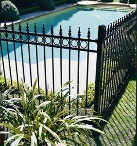 Black metal pool fence surrounding a rectangular pool with clear water. Green grass and plants are in the foreground.