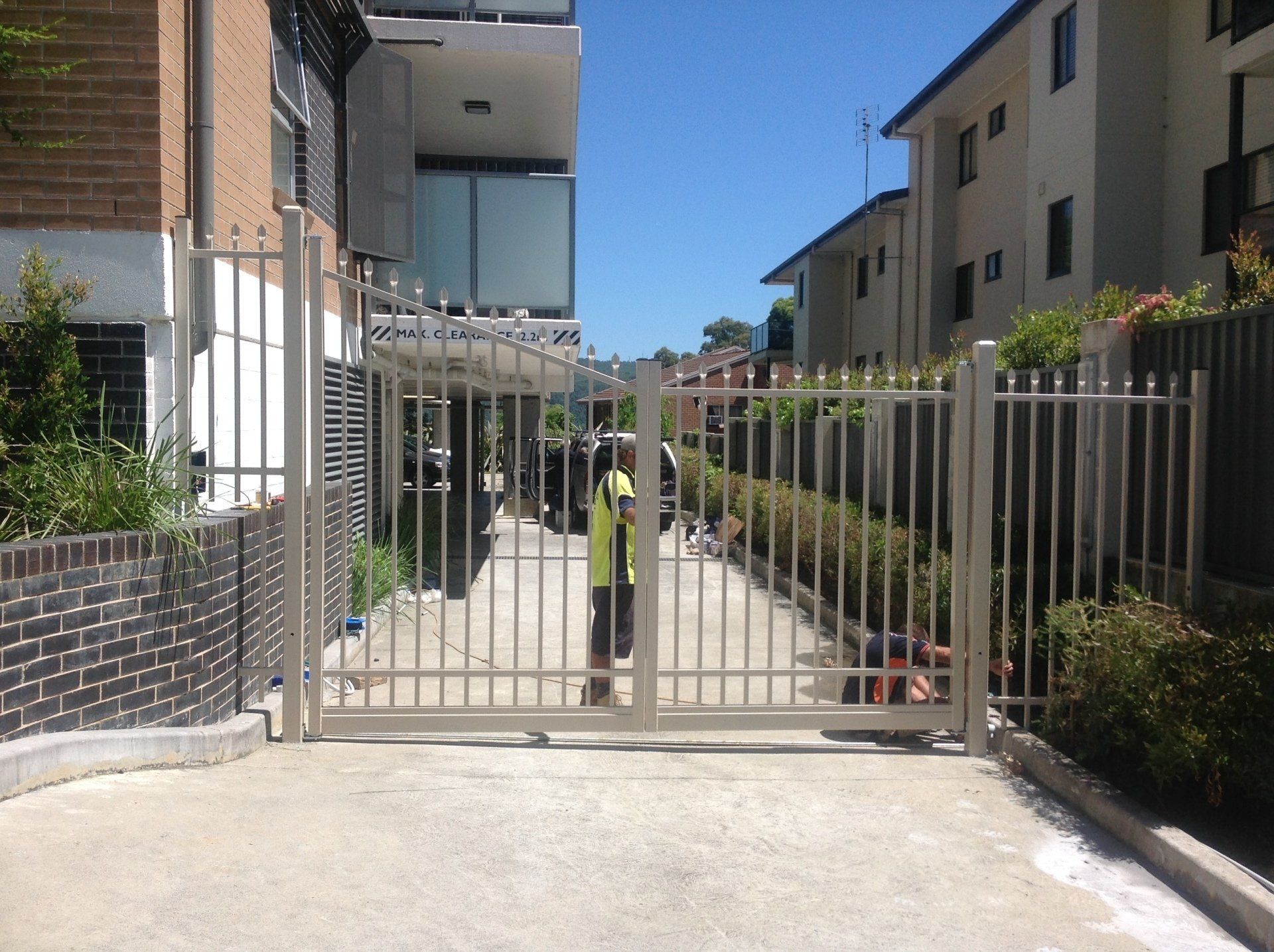 Metal gate opening into a driveway between two buildings. Person in yellow vest. Sunny day.