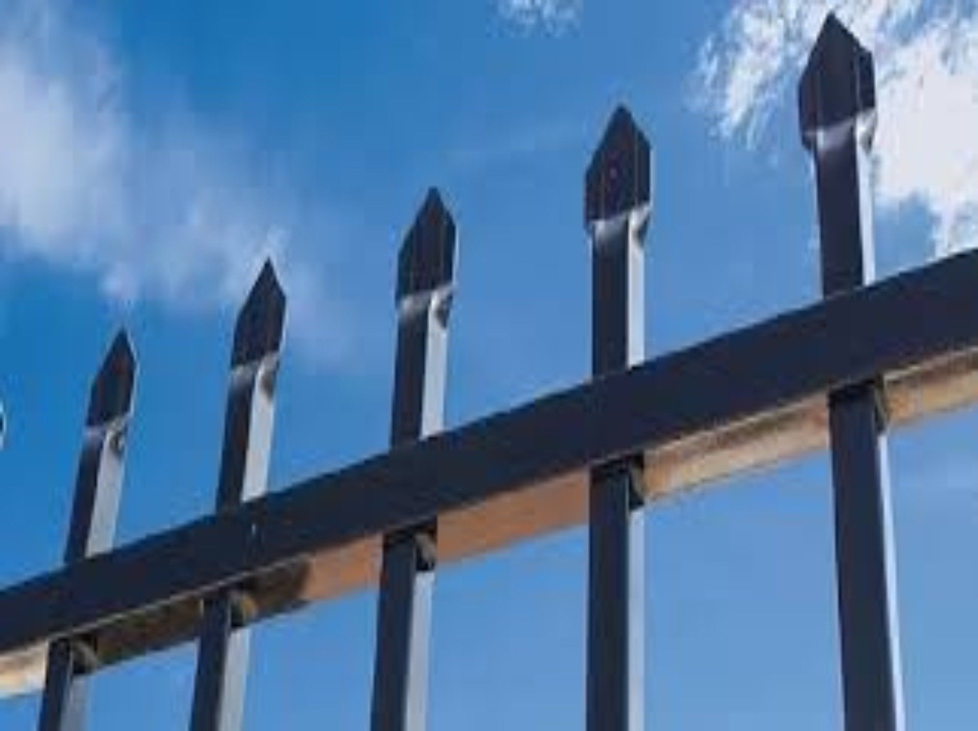 Black metal fence with pointed tops against a blue sky with clouds.