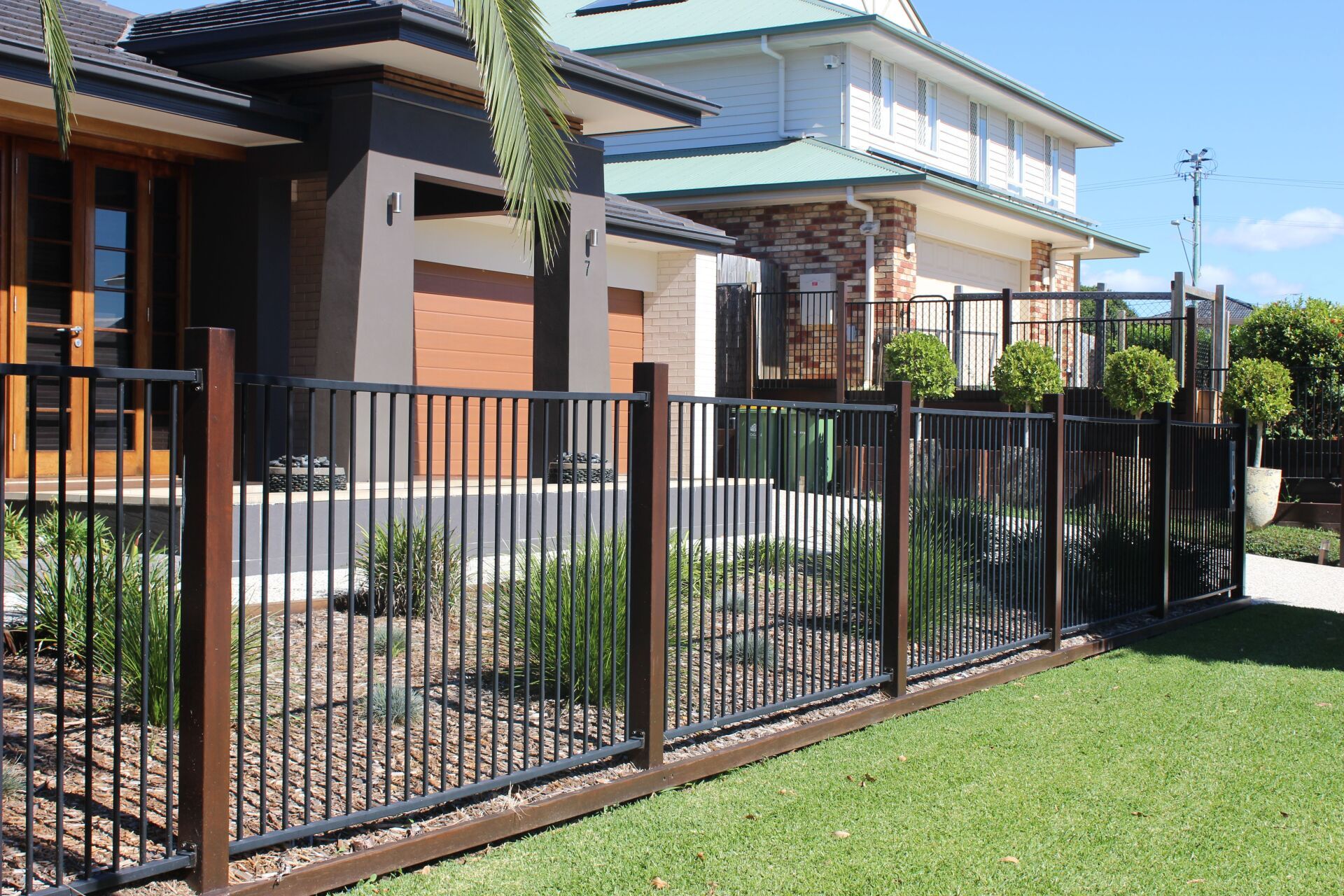 Black metal fence with wood posts in front of two houses. Green grass in foreground.