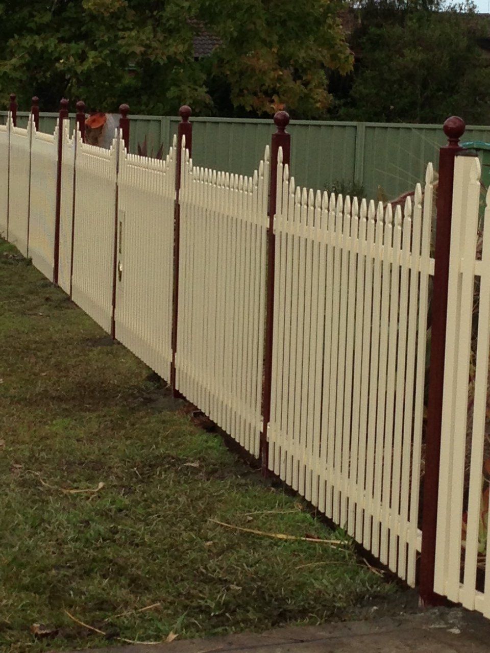 White picket fence with dark brown posts and caps, set against a green lawn and backdrop.