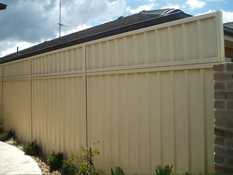 Beige corrugated metal fence next to a brick wall, with dark roof in background.