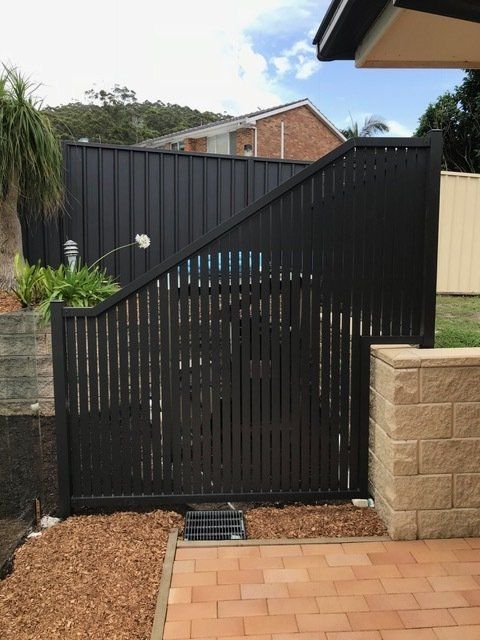 Black slatted fence with a matching solid section. Behind the fence is a house and trees.