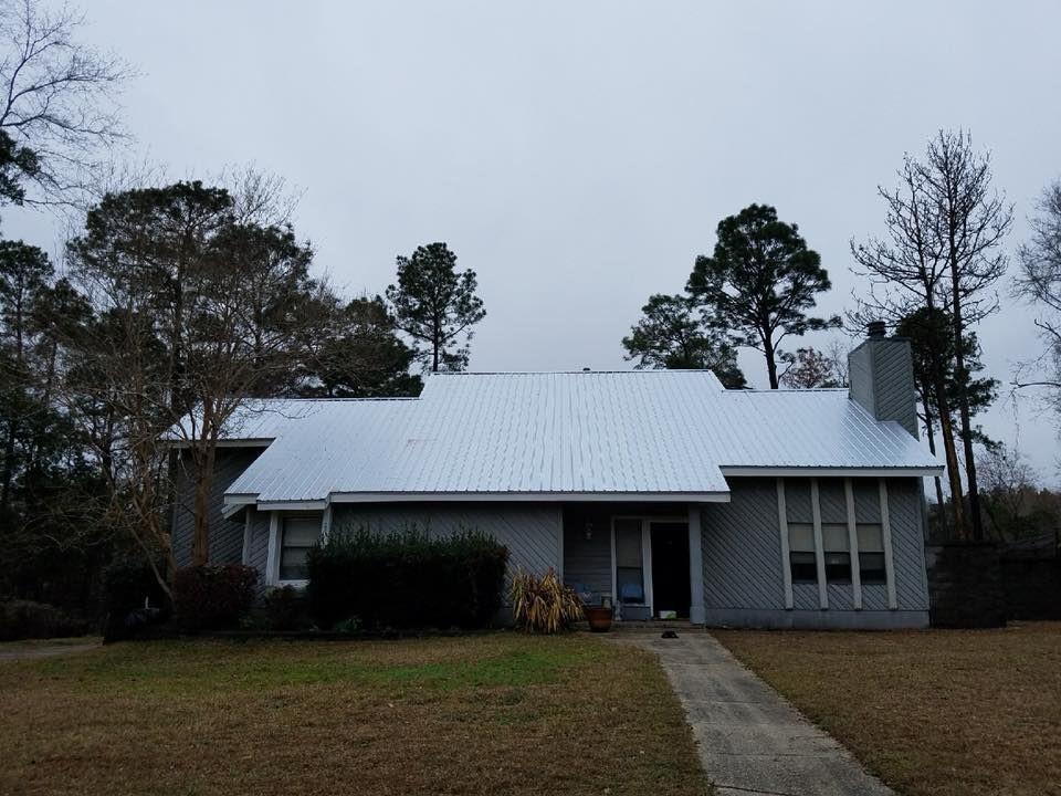 Gray house with metal roof on a cloudy day, surrounded by trees and a pathway leading to the door.