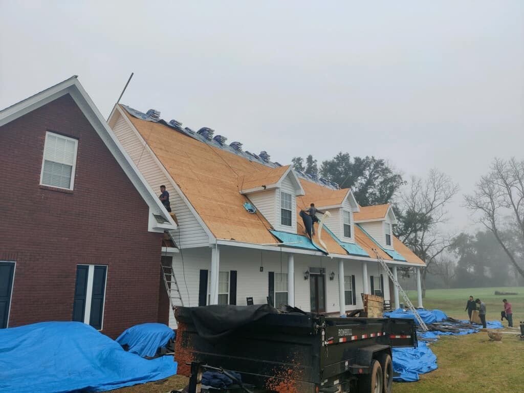 Roofers working on a house with a red brick side and white facade, surrounded by blue tarps and a trailer.