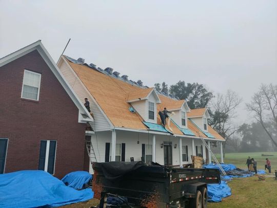 Roofers working on a house with a red brick side and white facade, surrounded by blue tarps and a trailer.