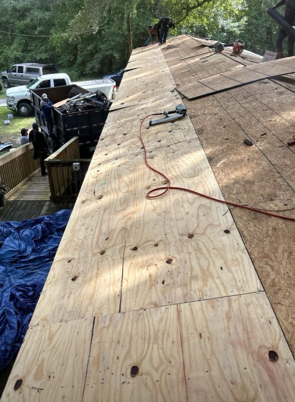 Roofers working on a house roof. Plywood sheets being installed. Trucks and debris visible.