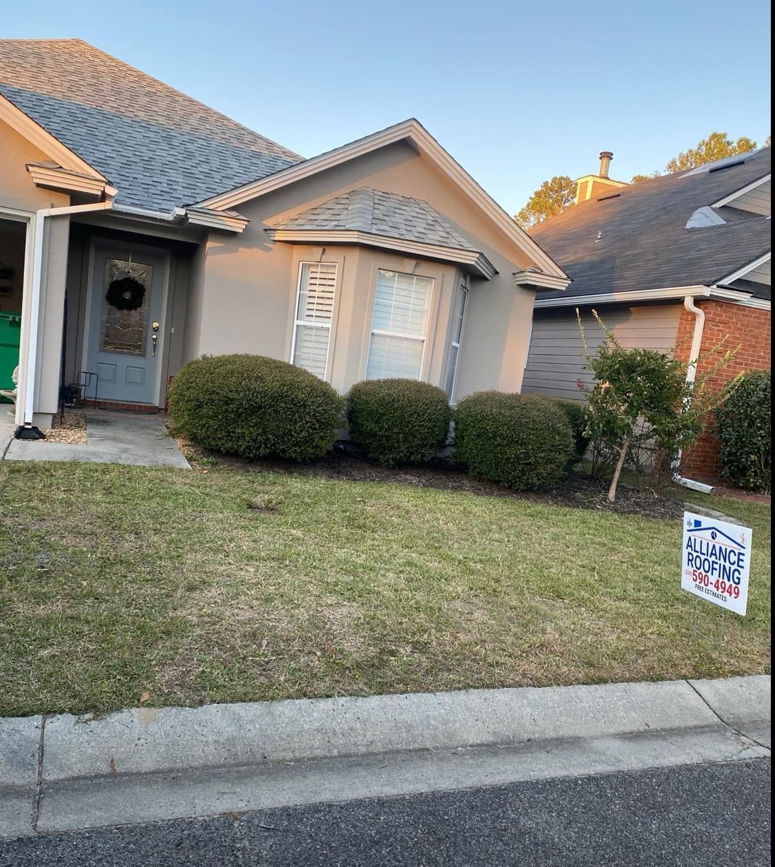 A house with a gray roof and light gray walls. Green bushes line the front, a lawn below.