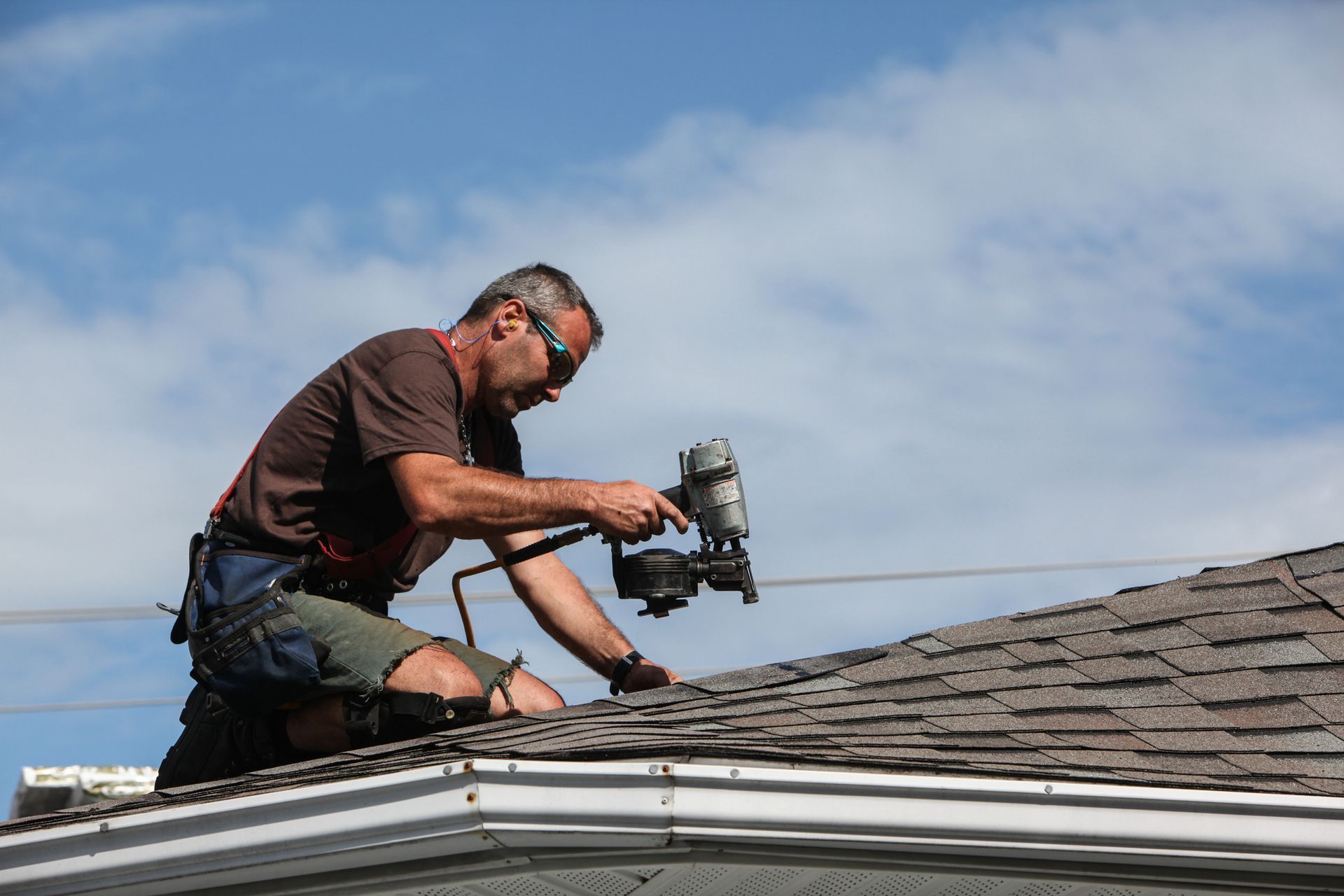Roofer on a roof uses a nail gun, wearing safety glasses and a harness, against a blue sky.