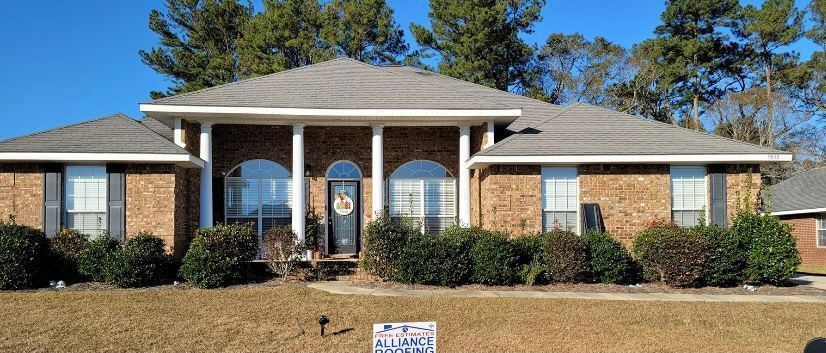 A brick home with a gray roof and black shutters, trees and blue sky visible.