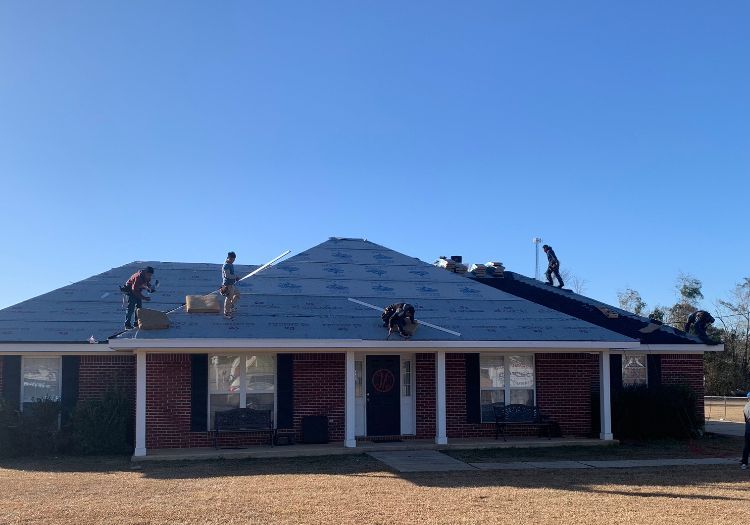Workers installing a roof on a brick house with blue shingles on a sunny day.