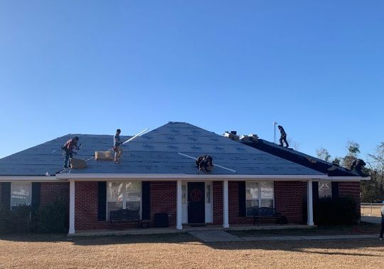 Workers installing a roof on a brick house with blue shingles on a sunny day.
