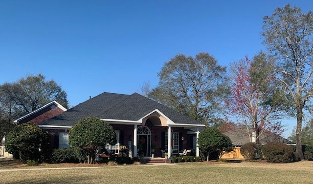House with dark roof, brick and white trim, shrubs, trees, and blue sky.