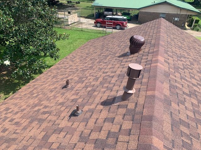Brown shingled roof with vents. A red vehicle and green roof visible in the background. Sunny day.