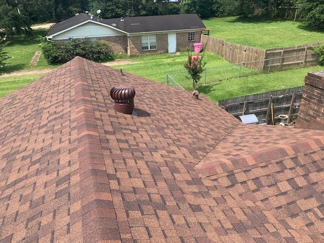 Brown shingled roof with a vent in the center, a brick chimney, and a grassy backyard with a fence.