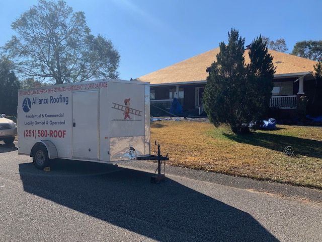 White trailer for Alliance Roofing parked on a street with a house under roof repair in the background.