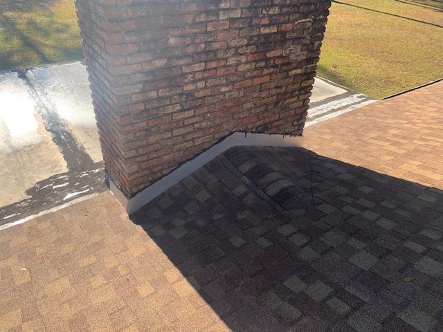 Brick chimney on a shingled roof with flashing, casting a shadow on the shingles.