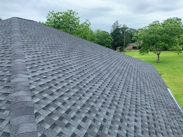 Dark gray asphalt shingle roof on a house, angled view, green trees in background.
