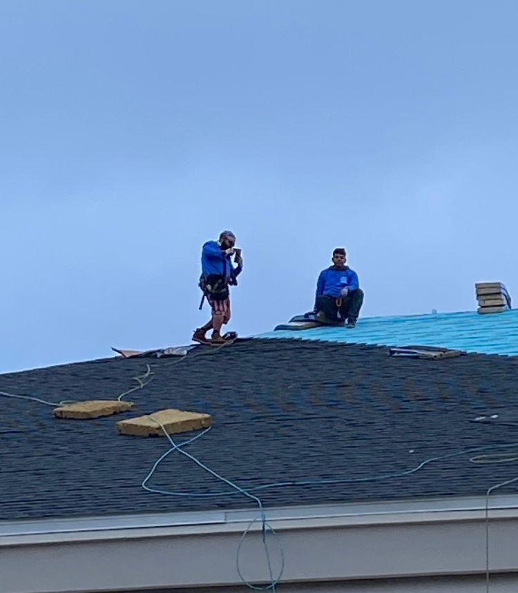 Two roofers in blue shirts on a roof, installing shingles. One is standing, the other is seated.
