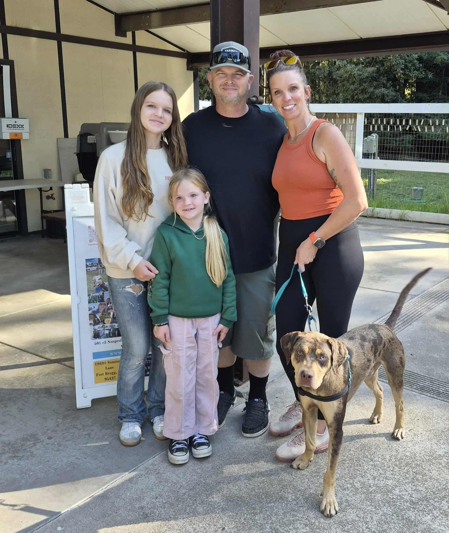 Family of four with a dog, posing outdoors by a building; dog is on a leash.