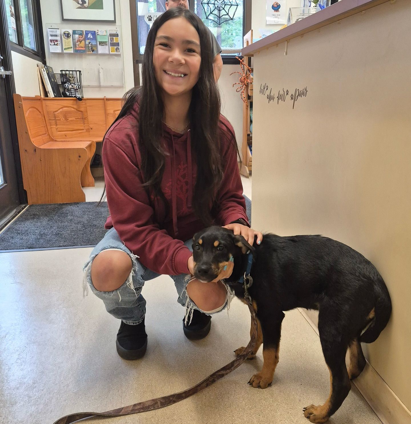 Young person kneels with a black and tan dog in a room. Smiling person in ripped jeans pets the dog.