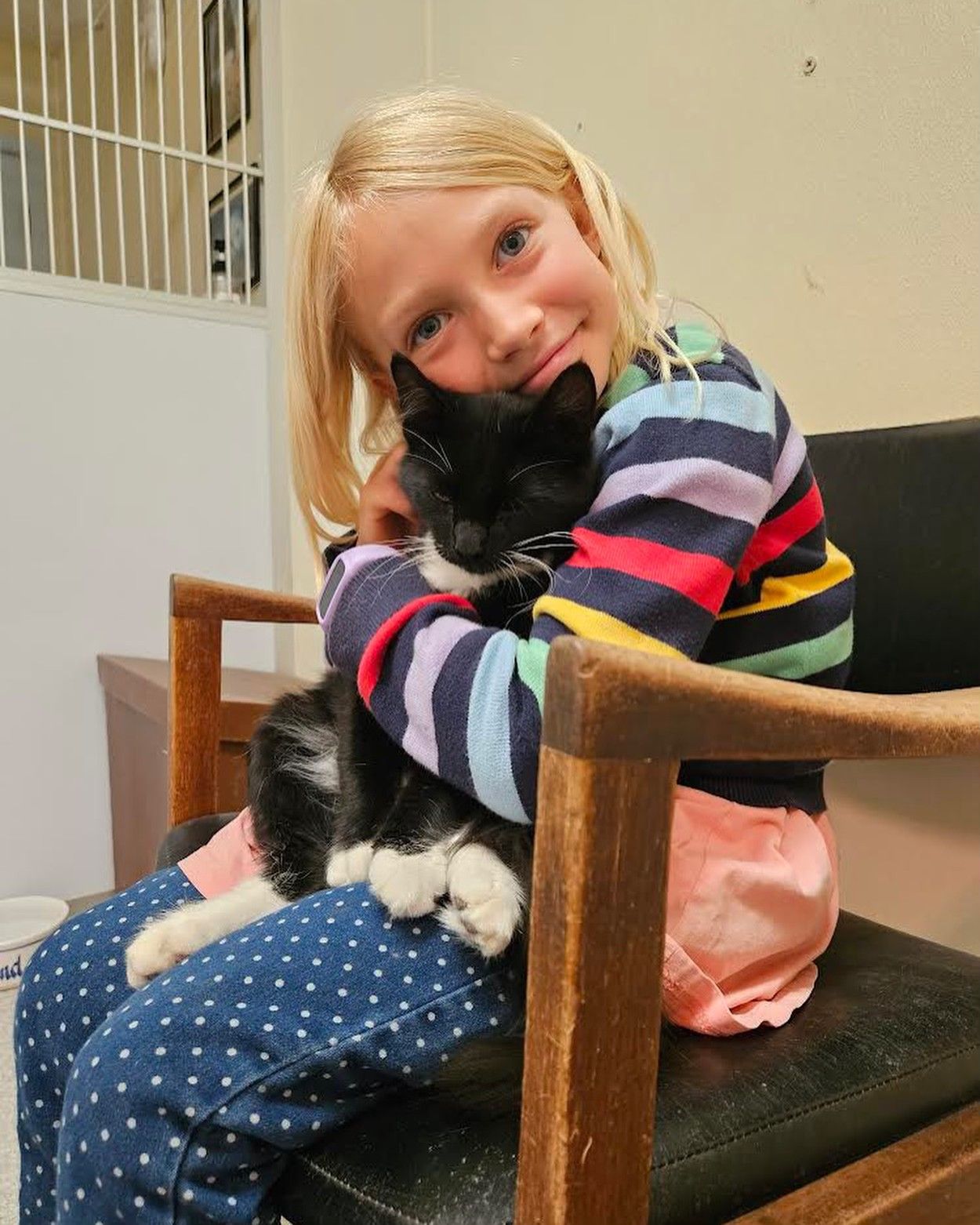 Girl in colorful striped shirt cuddles a black and white cat on her lap in a waiting room.
