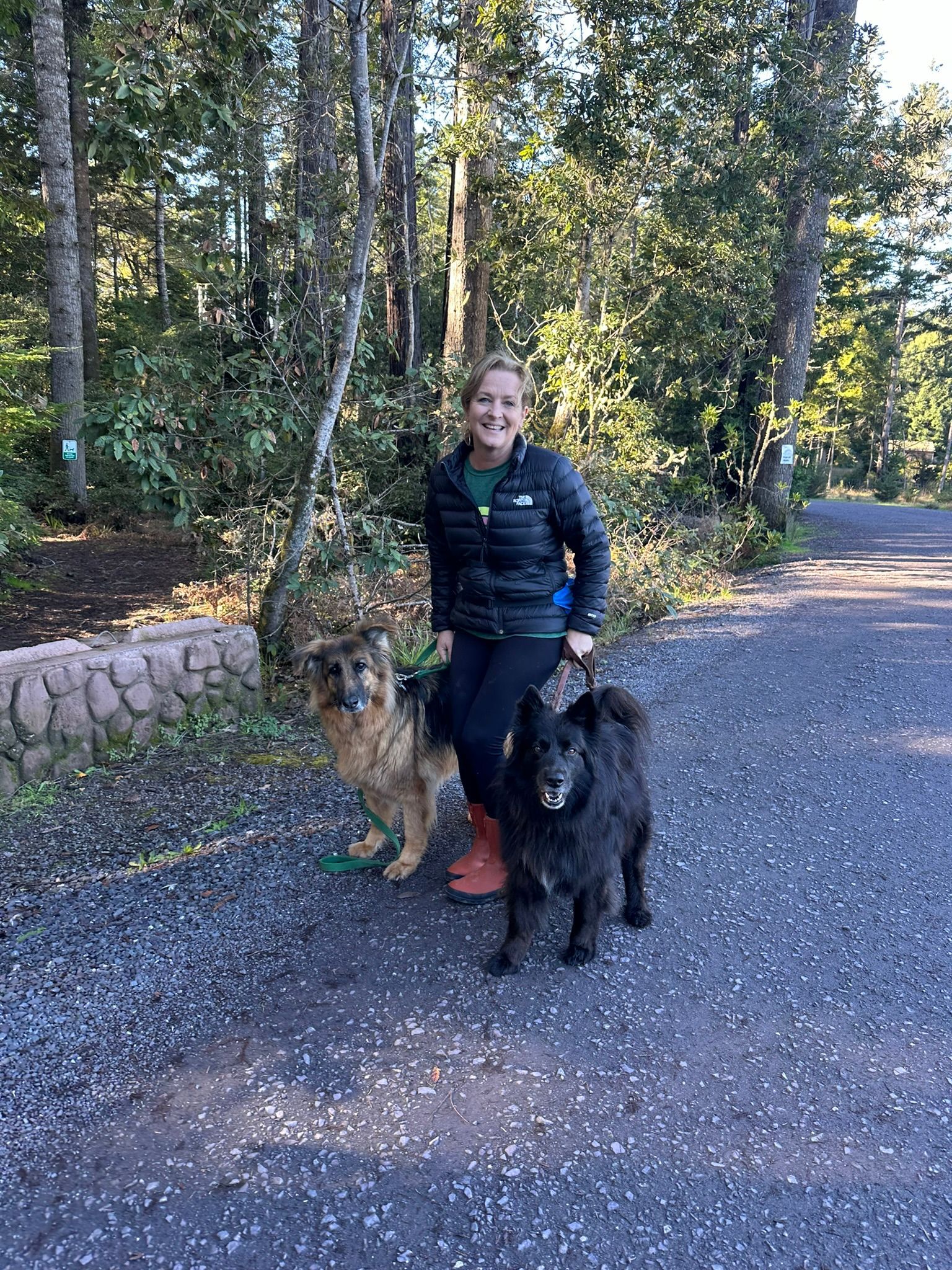 Woman smiling with two dogs on a gravel path near trees.
