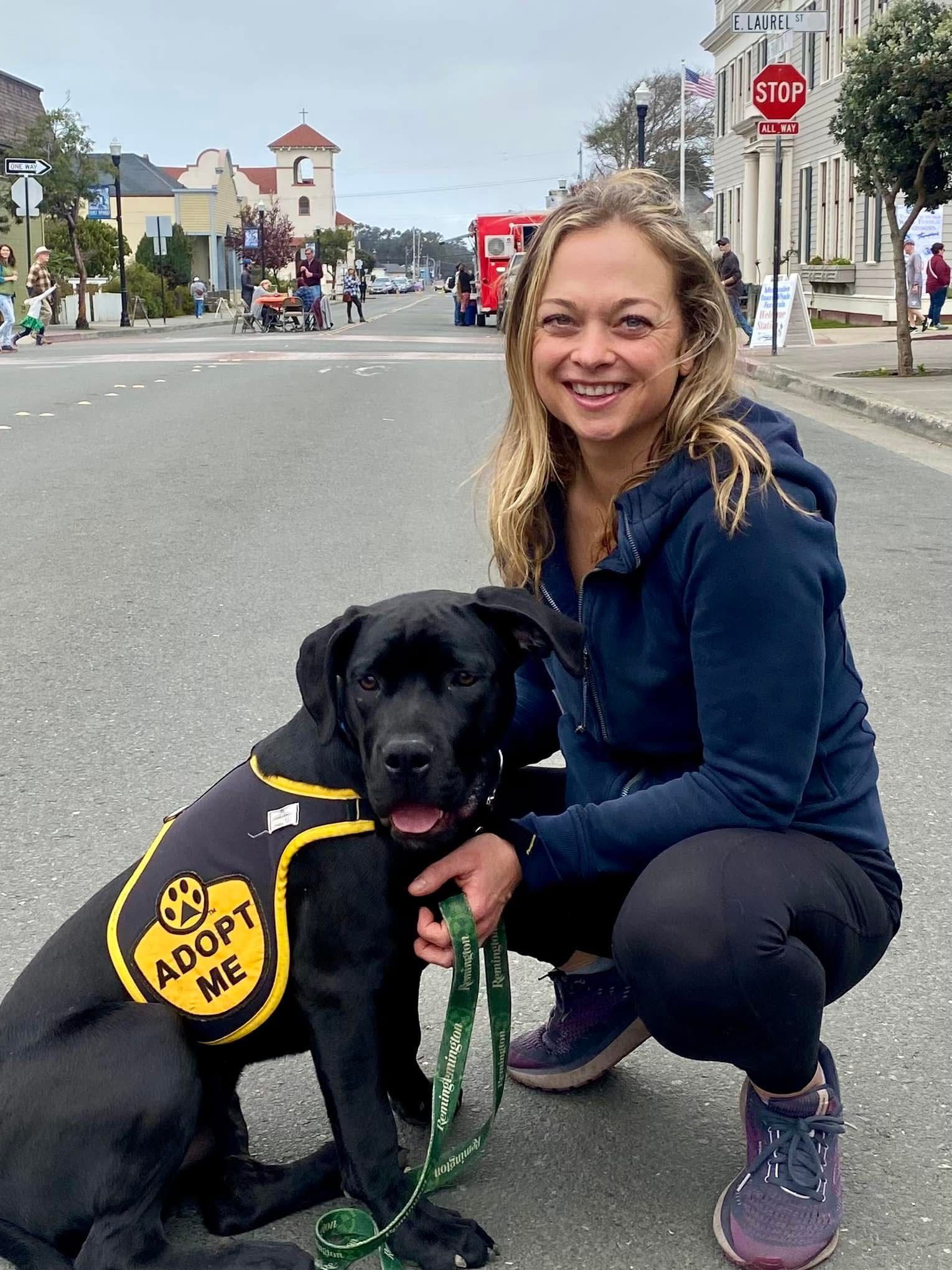 Woman crouches next to a black dog wearing a 