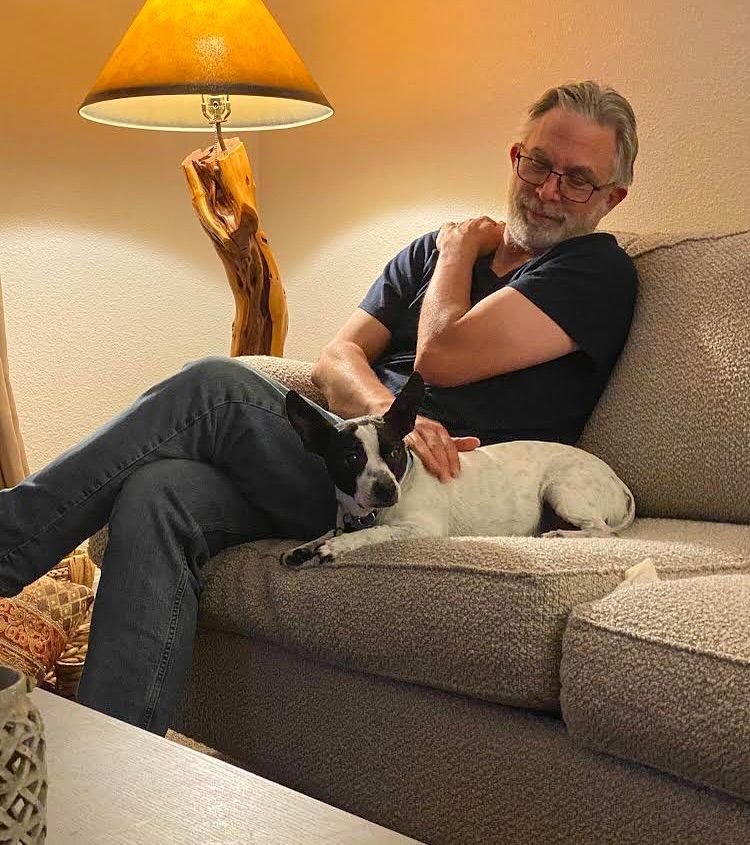 Man with glasses on couch, petting small black and white dog. Lit by a lamp.