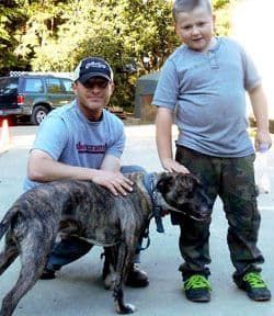 Man and boy petting a brindle dog outdoors. Boy in camo pants, man in cap.