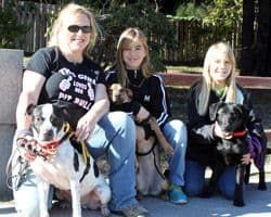 Three women and two dogs posing outside; one dog is on a leash.