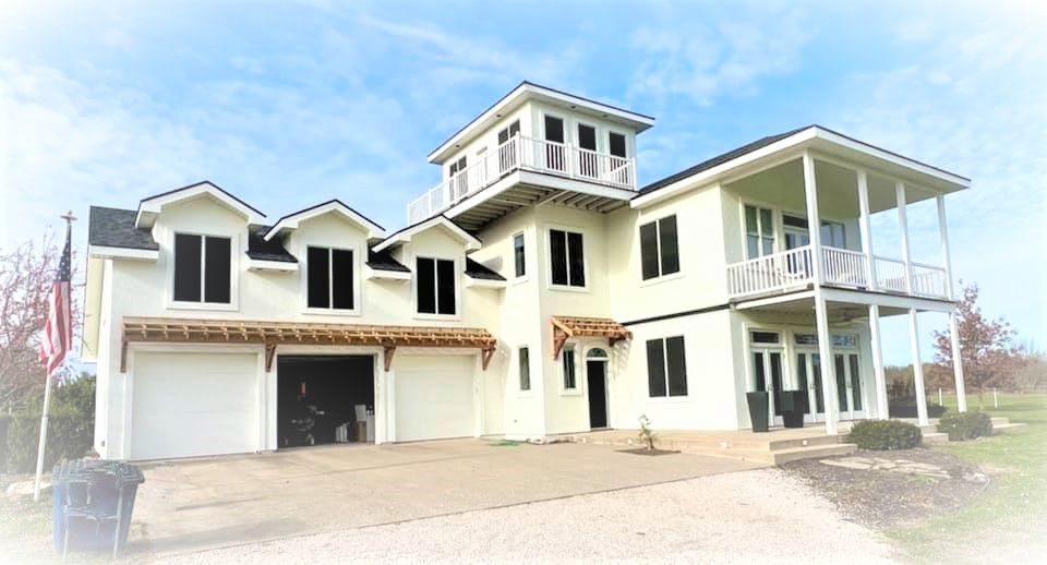 Two-story beige house with a tower and balcony, set on a gravel driveway under a blue sky.