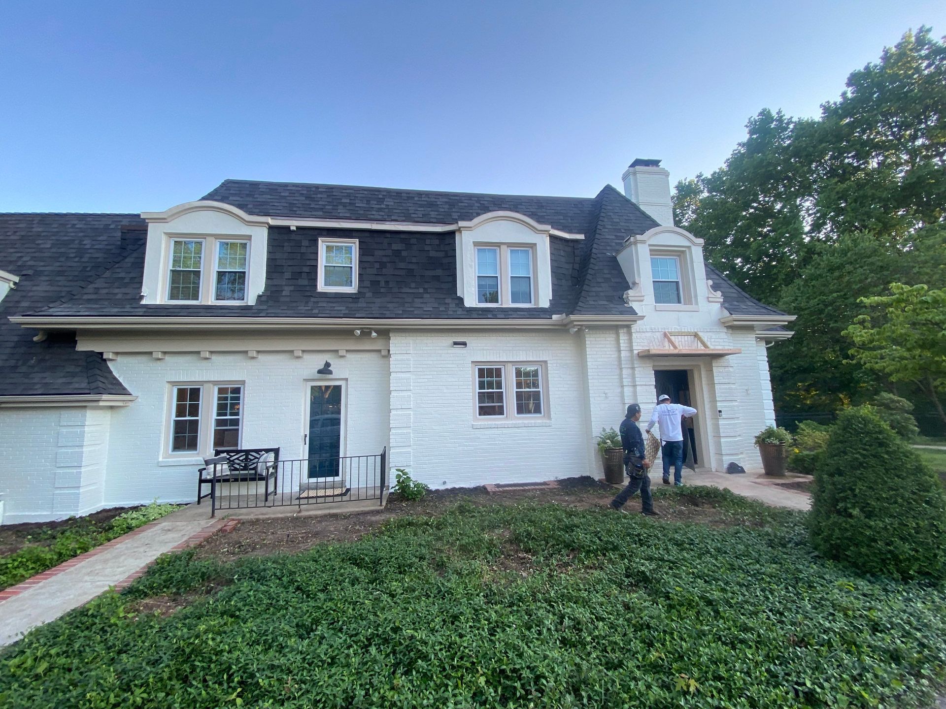 White house with black roof, two people entering doorway, landscaping in front.