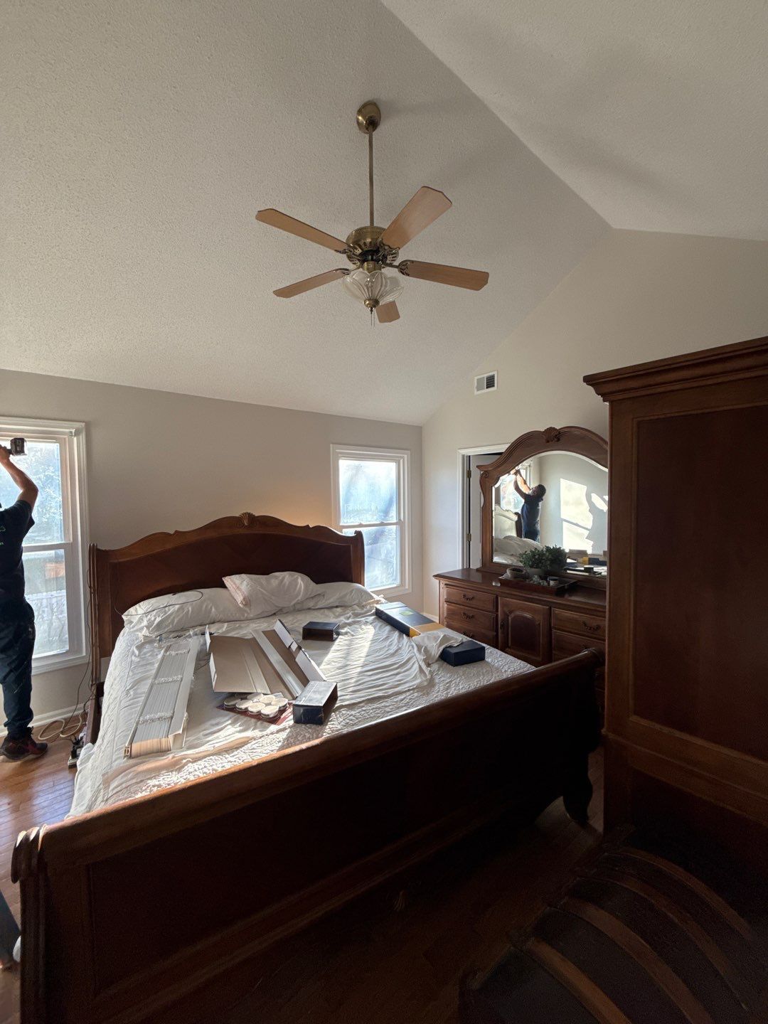 Bedroom with wooden furniture, ceiling fan, and person working by the window.