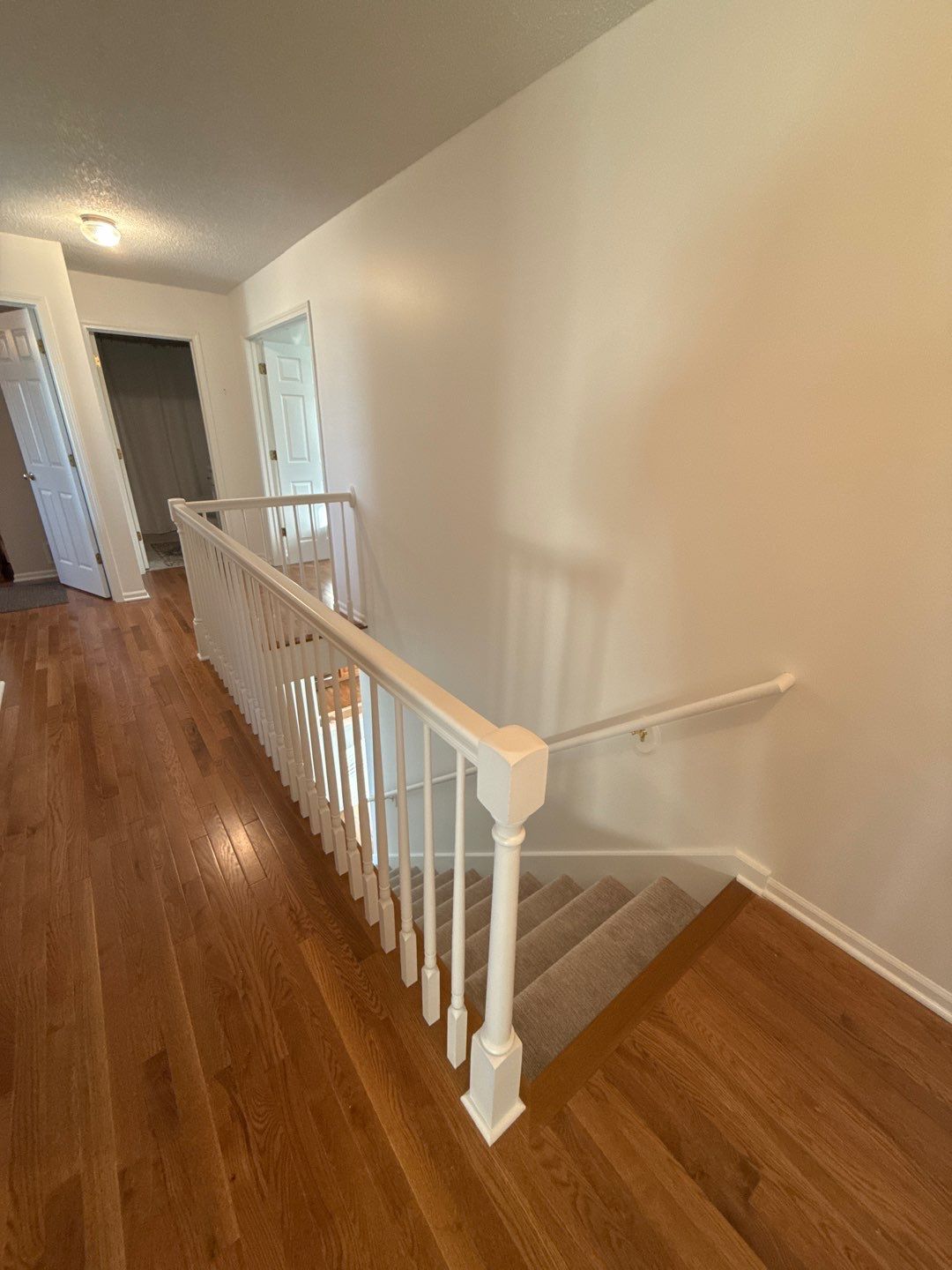 Top-down view of a hallway with white railings and stairs leading down. Light wood flooring and white walls.