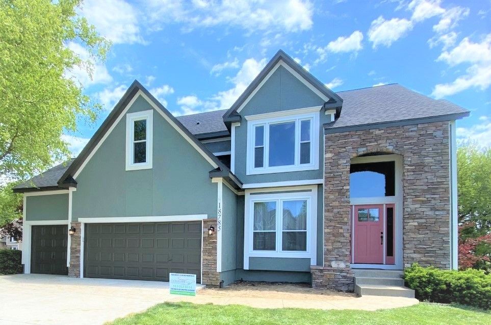Two-story house with blue-green siding, stone facade, and a pink front door under a bright sky.