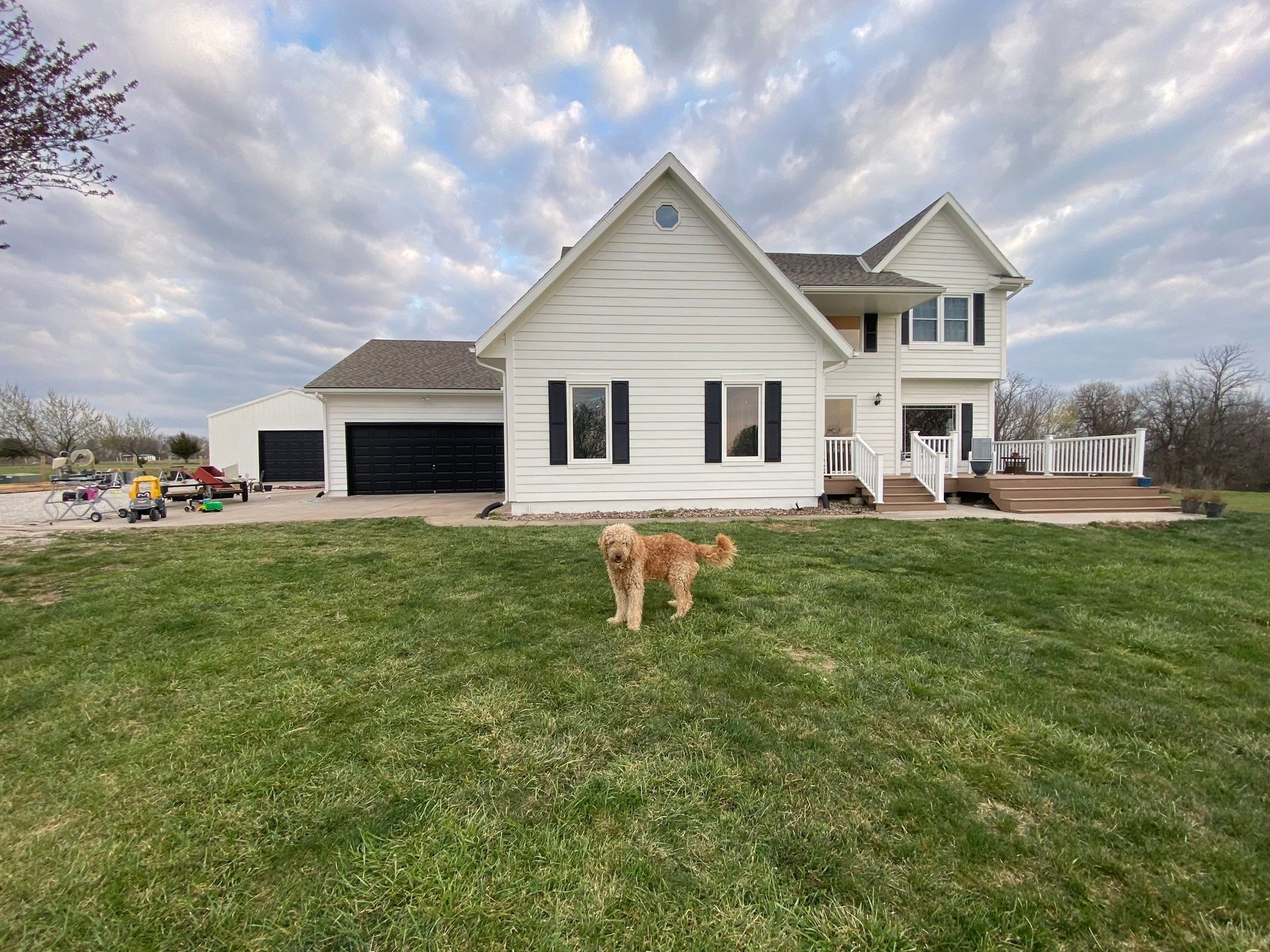A dog stands in front of a white house with black garage doors, on a green lawn under a cloudy sky.