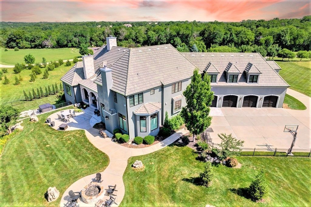 Large, blue-gray home with gray roof, attached garage, and manicured lawn. Trees and sky in the background.