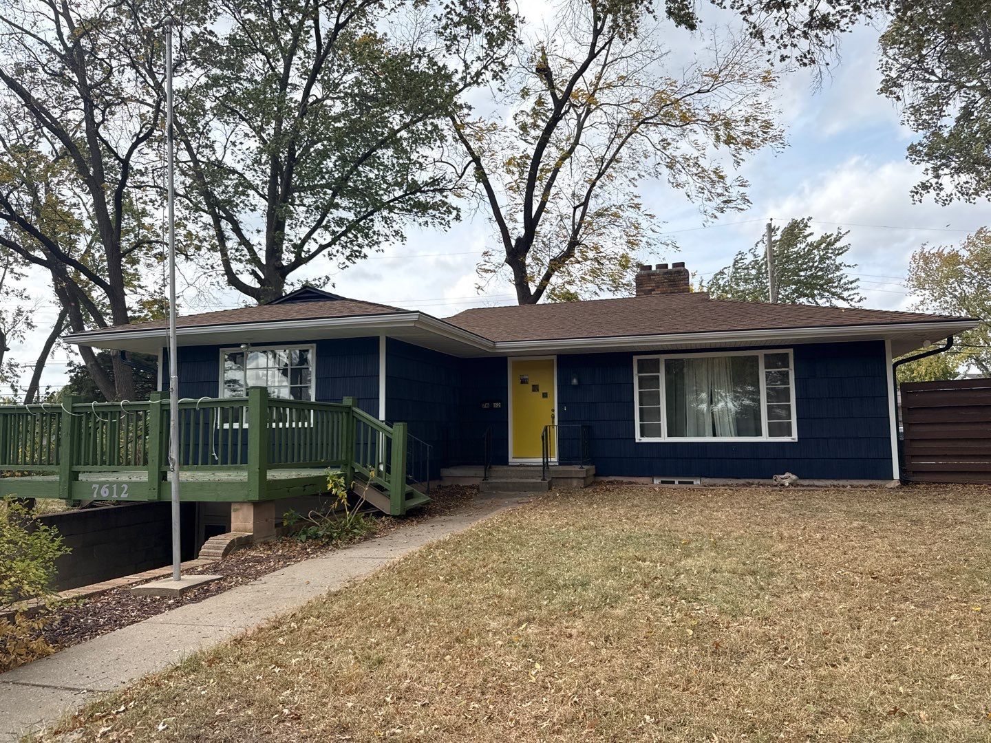 Dark blue house with yellow door, brown roof, green deck, and brown yard.