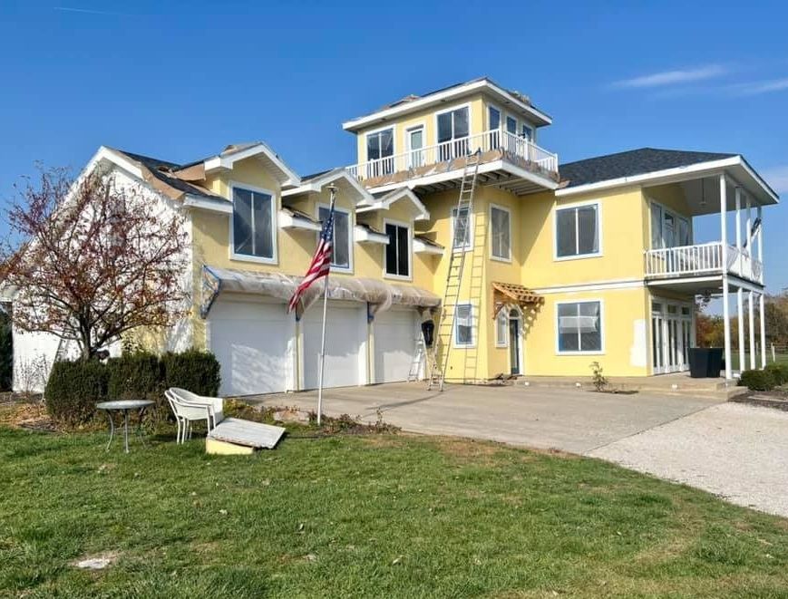 Yellow house with garage, balconies, and tower under construction; American flag in front.