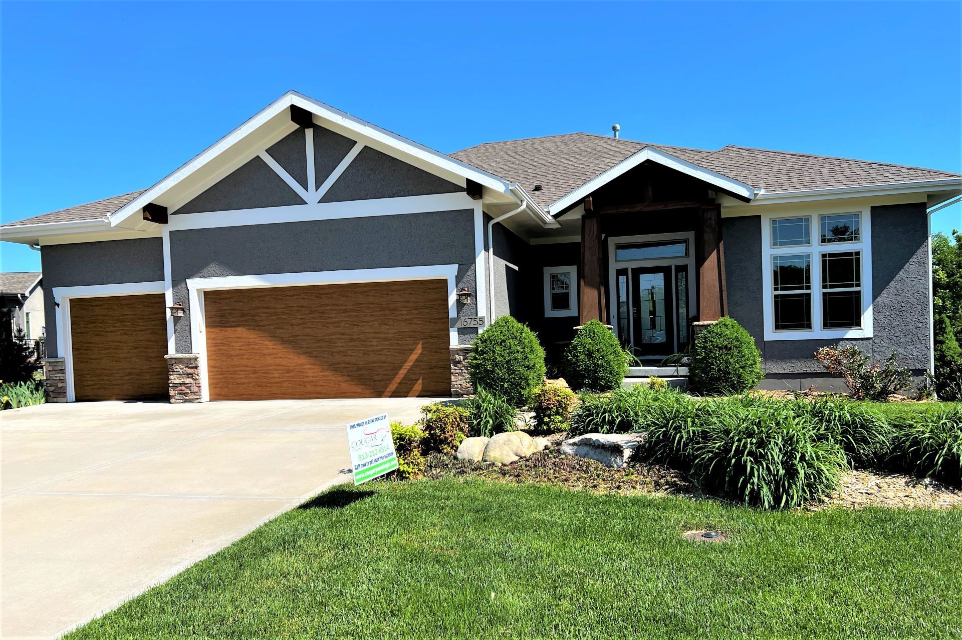Gray stucco house with brown garage doors, landscaped front yard, clear blue sky.