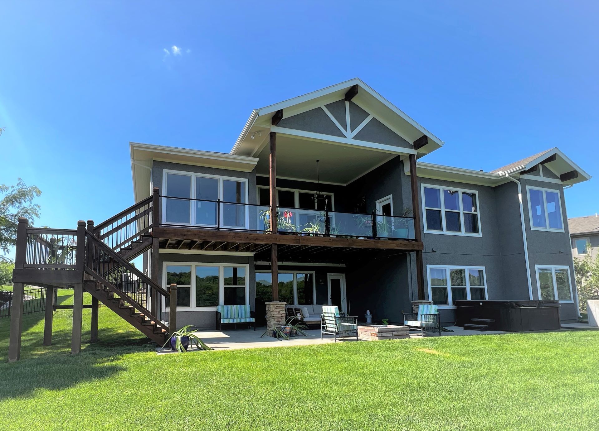 Two-story gray house with a wooden deck and glass railings. Sunny day with a clear blue sky.