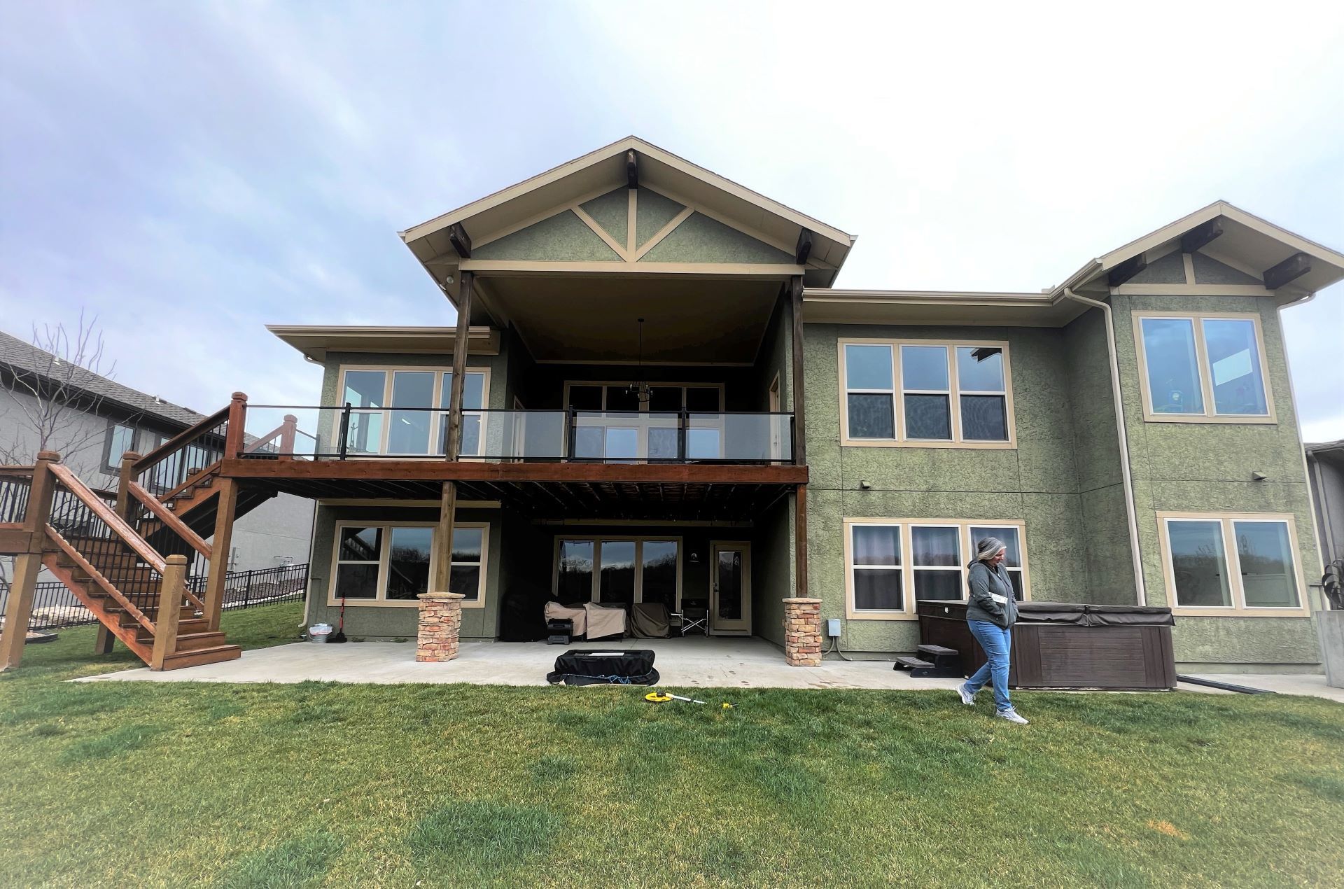Back of a two-story house with a wooden deck, glass railings, and a person standing near a hot tub on the lawn.